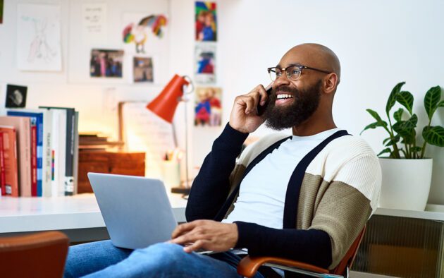 A man with glasses and a beard smiles while talking on a phone and using a laptop in a brightly-lit office with books and plants around him.