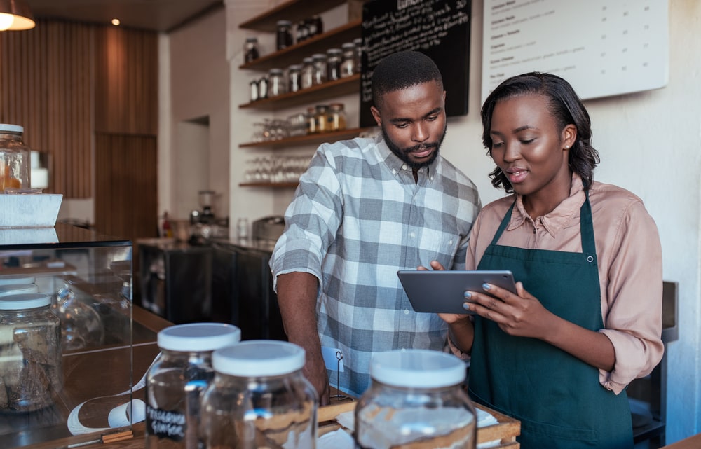 Two people in a cafe, one wearing an apron, looking at a tablet. Shelves with jars are in the background.