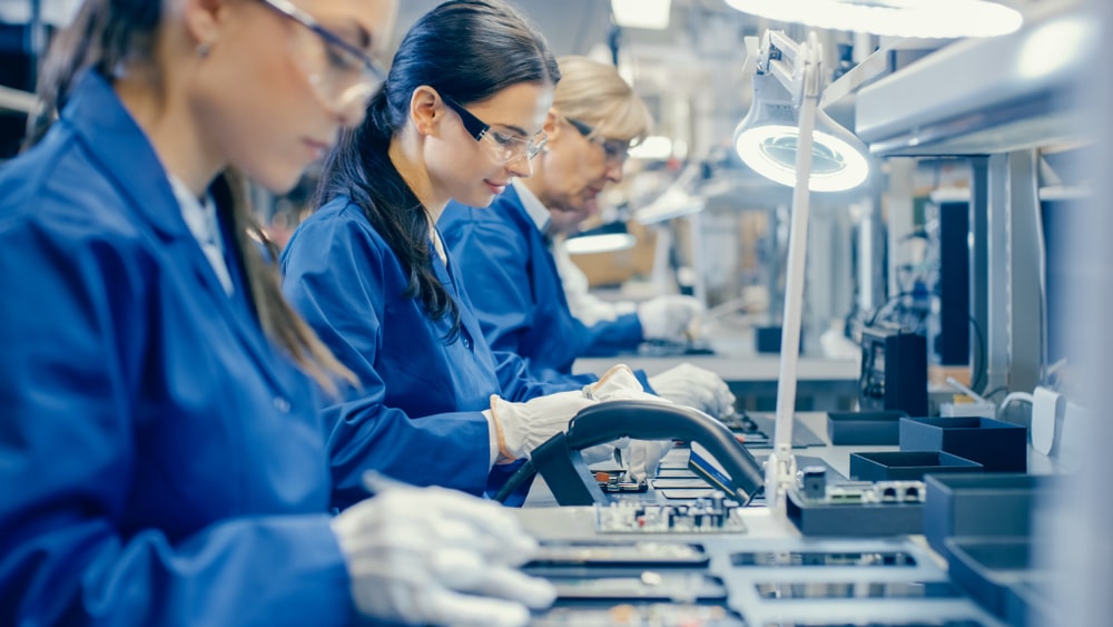 People in blue lab coats and safety glasses work at a table assembling electronic components in a well-lit industrial setting.