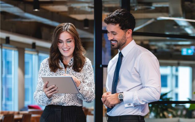 Two people in business attire stand in an office. One holds a tablet, and they both appear engaged in conversation.