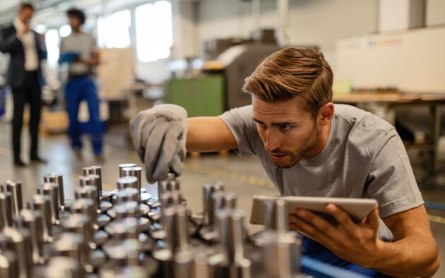 A man inspects metal components on a table in a factory setting while holding a tablet. Another person stands in the background.