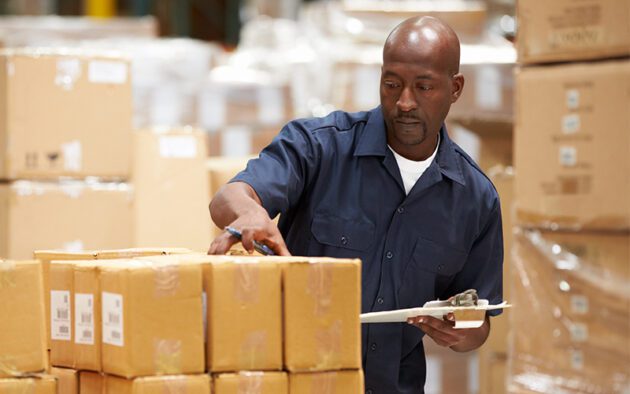 Man in a warehouse checking inventory, holding a clipboard, surrounded by stacked boxes.