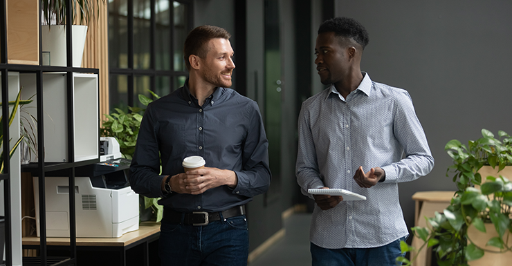 Two men walking and talking in an office. One holds a coffee cup, the other a notebook. They are surrounded by plants and office furniture.