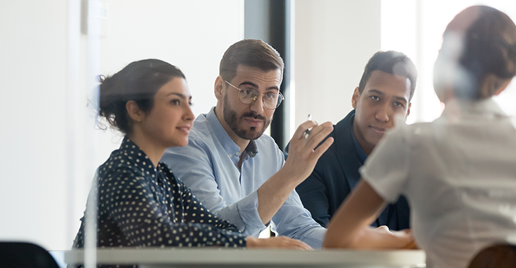 Three people in business attire sit at a table, attentively listening and engaging with a person across from them in an office setting.