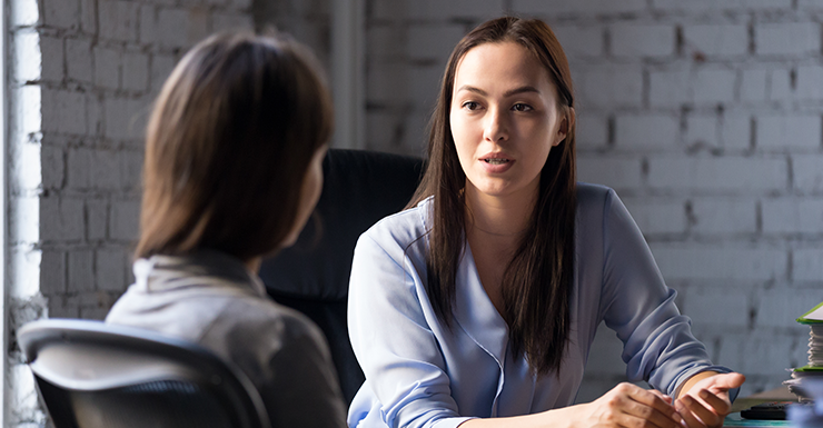 Two women are engaged in a conversation in an office setting, with one facing the camera and the other with her back turned. They are seated at a table against a white brick wall.