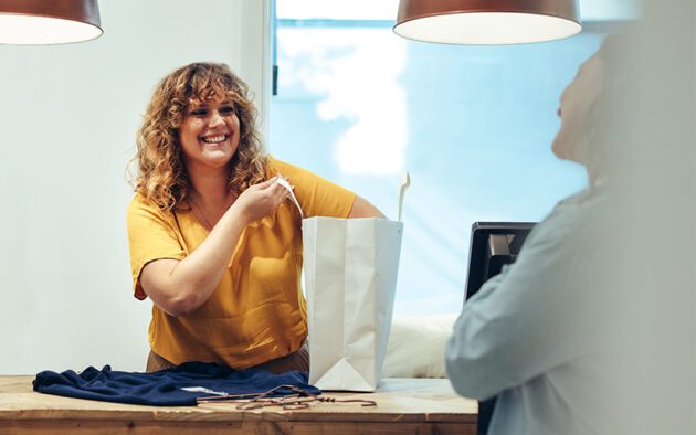 A woman in a yellow shirt smiles while placing items into a white shopping bag at a counter, engaging with another person.