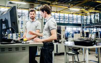 Two people in a factory setting stand by a desk with computers, engaged in conversation. The industrial backdrop includes machinery and large windows.