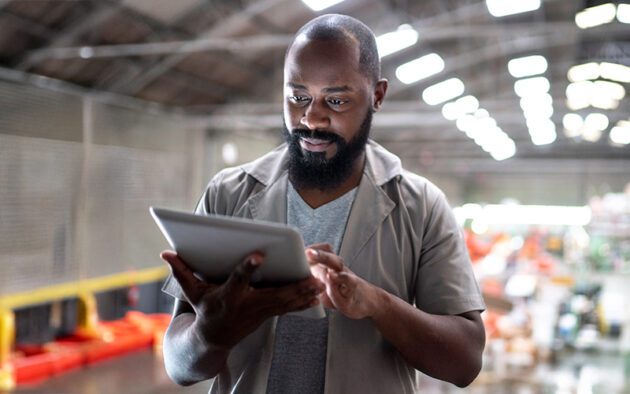 A man stands in a warehouse using a tablet, with blurred shelves and equipment visible in the background.