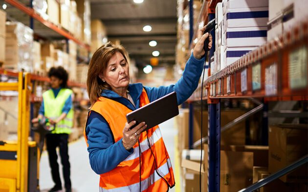 A woman in an orange vest uses a handheld scanner and tablet to check inventory on warehouse shelves. Another worker is visible in the background.