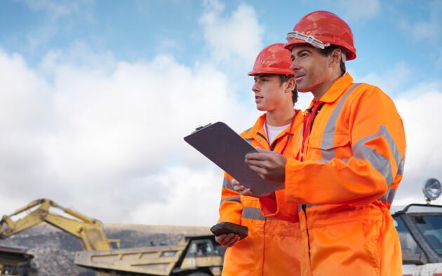 Two construction workers in orange safety gear and helmets stand outdoors, reviewing a mining report with a clipboard and phone in hand, as construction vehicles rumble in the background.