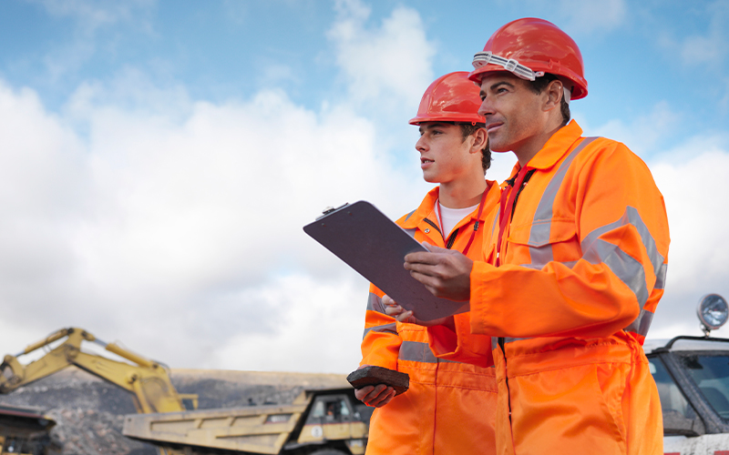 Two construction workers in orange safety gear and helmets stand outdoors, reviewing a mining report with a clipboard and phone in hand, as construction vehicles rumble in the background.