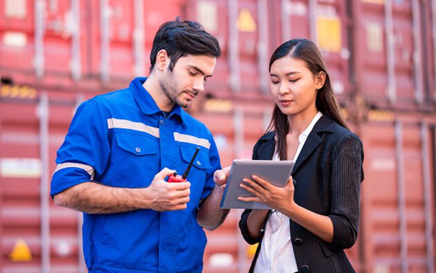 Two people stand in front of shipping containers. One wears a blue work uniform and holds a radio, while the other in a blazer uses a tablet.