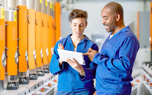 Two workers in blue uniforms engage in a discussion about employee experience while examining a tablet amidst the vibrant orange machinery of the factory.