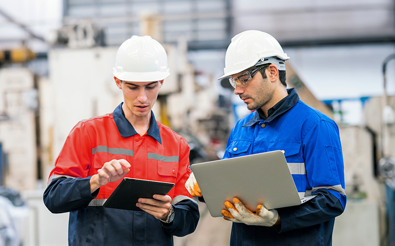 Two industrial workers in safety helmets and protective clothing review information on a tablet and a laptop inside a factory setting.