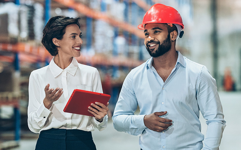 A woman holding a tablet talks with a man wearing a red hard hat in a warehouse setting.