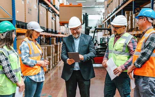 A group of people in safety helmets and vests gather around a person holding a tablet in a warehouse setting.