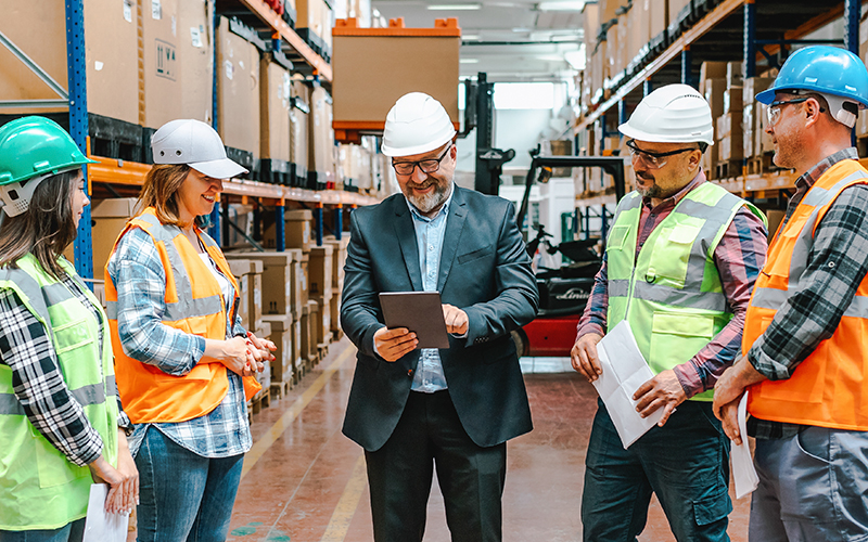 A group of people in safety helmets and vests gather around a person holding a tablet in a warehouse setting.