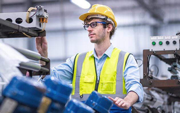 A man in a yellow safety vest and hard hat operates machinery in an industrial setting.