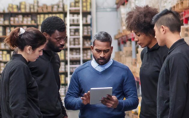 A group of five people in a warehouse gather around a person holding a tablet, discussing work. Shelves filled with boxes are visible in the background.