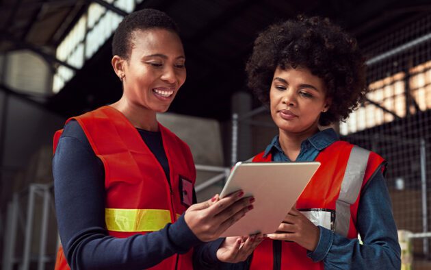 Two people wearing red safety vests stand indoors, looking at a tablet device together. One person is smiling while showing the tablet to the other.