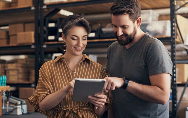 Two people stand in a warehouse, looking at a tablet and smiling. Shelves with boxes are visible in the background.