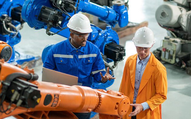 Two engineers wearing helmets and protective clothing discuss near industrial robots in a factory setting.