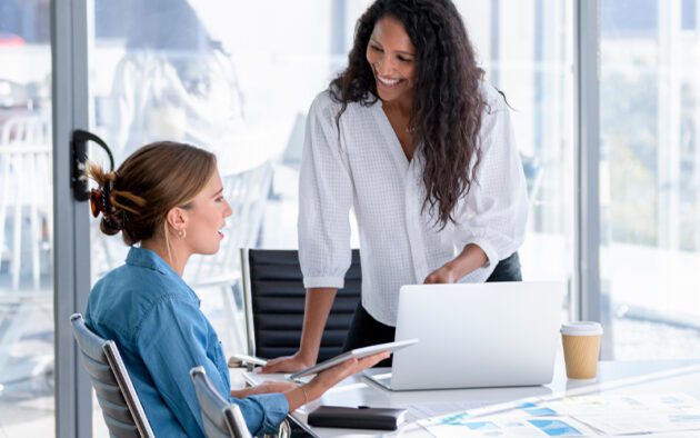 Two women in an office setting, one seated with a tablet and papers, the other standing and smiling beside a laptop. A coffee cup is on the table.