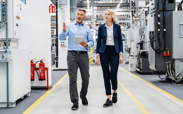 Two people in business attire walk and talk inside a modern industrial facility, surrounded by machinery and equipment. One person holds a tablet.