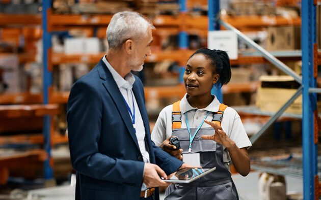 In a bustling warehouse, two individuals confer amidst looming shelves stacked with boxes. One holds a tablet, ensuring every detail aligns with compliance standards, while the other, dressed in a work uniform with a walkie-talkie clipped on, nods in understanding as they discuss logistics.