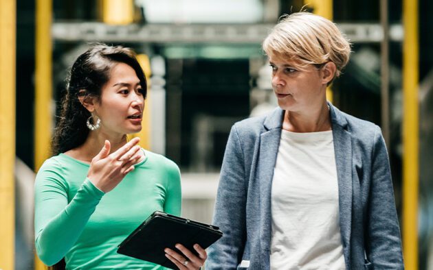 Two people are engaged in conversation indoors, with one holding a tablet. They appear to be discussing something work-related.