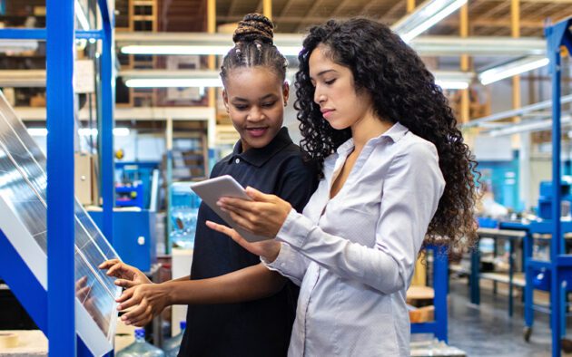 Two women in a factory setting, one holding a tablet and the other interacting with machinery, both focused on their tasks.