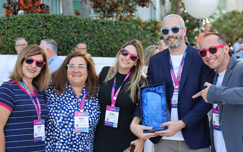 Five people wearing name tags and red sunglasses smile outdoors. One person holds a blue gift bag.