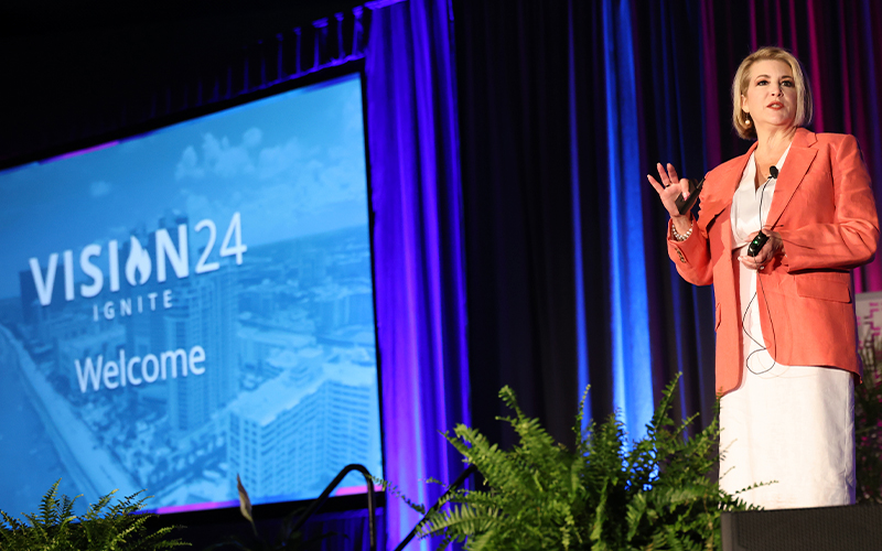A speaker in a coral blazer presents on stage at the Vision 24 Ignite event, with a "Welcome" slide displayed in the background.