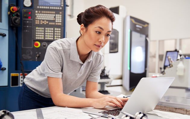 A woman in a grey polo shirt works at a laptop in an industrial setting with machinery and control panels in the background.