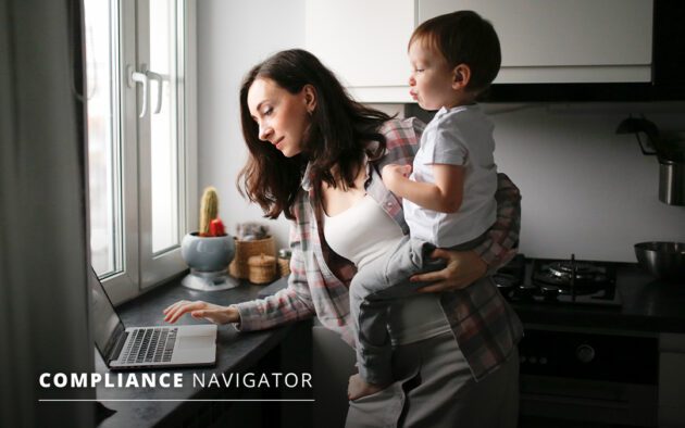 Woman holding child while working on a laptop in a kitchen