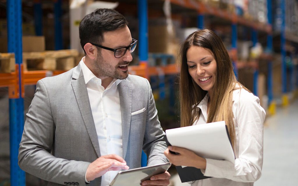Two office workers in business attire, standing in a warehouse, looking at a paper they are holding. Text overlays the image reads “Disruptions, Solutions, and Client Satisfaction: The Argument for Intelligent Workforce Management”. By WorkForce Software and Oracle