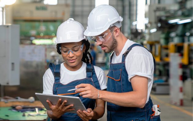 Two workers in a manufacturing facility, wearing hard hats, both looking at a tablet.