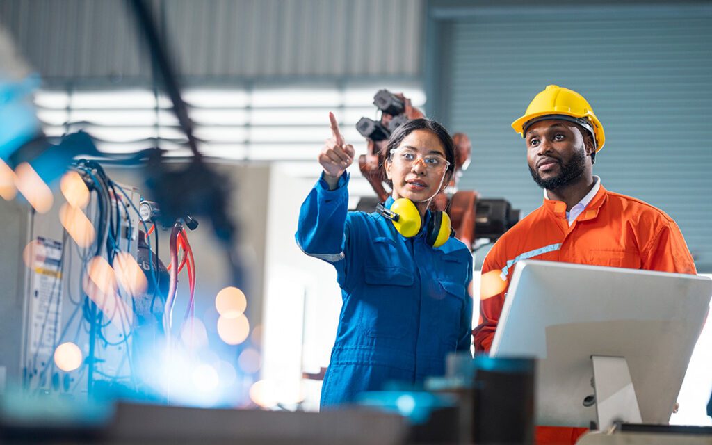 Two workers in a warehouse facility, wearing hard hats, standing in front of a computer monitor, both looking into the distance.