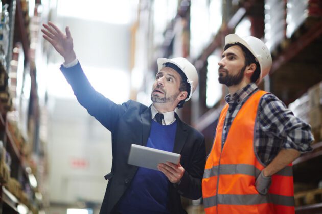 Two men in a warehouse with hard hats, one is wearing a safety vest while the other is wearing a suite and pointing at something above them.