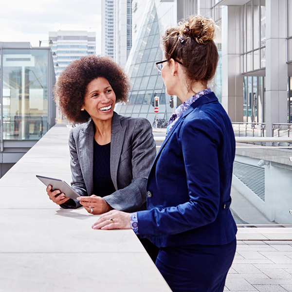 Two women in business attire are talking outside on an urban balcony, with modern buildings in the background. One woman holds a tablet, and both appear engaged in conversation.