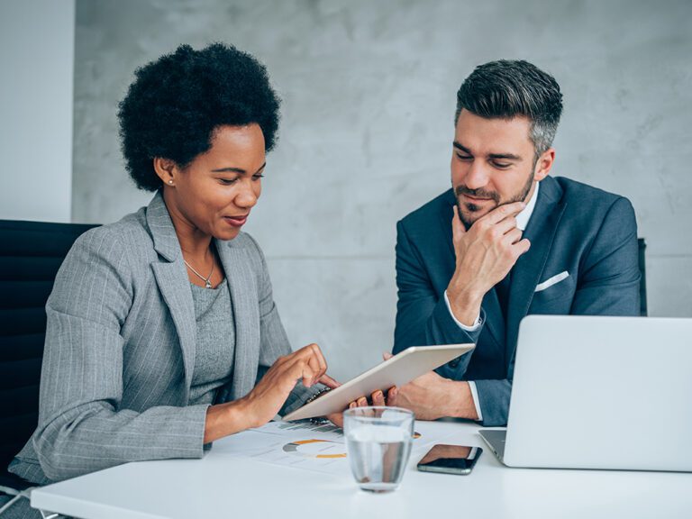 Two professionals in business attire discuss work over a tablet at a desk with a laptop, documents, and a glass of water.