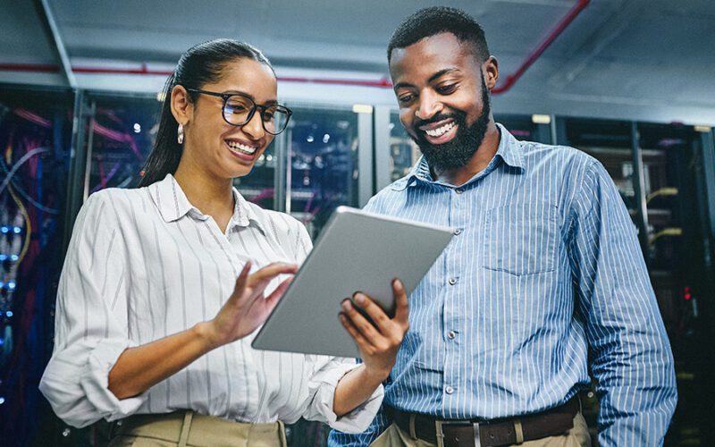 Two colleagues stand in a server room, smiling and looking at a tablet. The background consists of computer servers and cables.