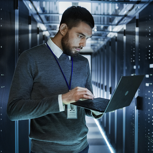 A man stands in a server room working on a laptop, wearing a grey sweater and ID badge. The background features rows of illuminated server racks.