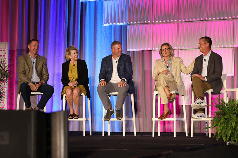 Five people sitting on tall chairs engage in conversation on a stage with blue and pink lighting.