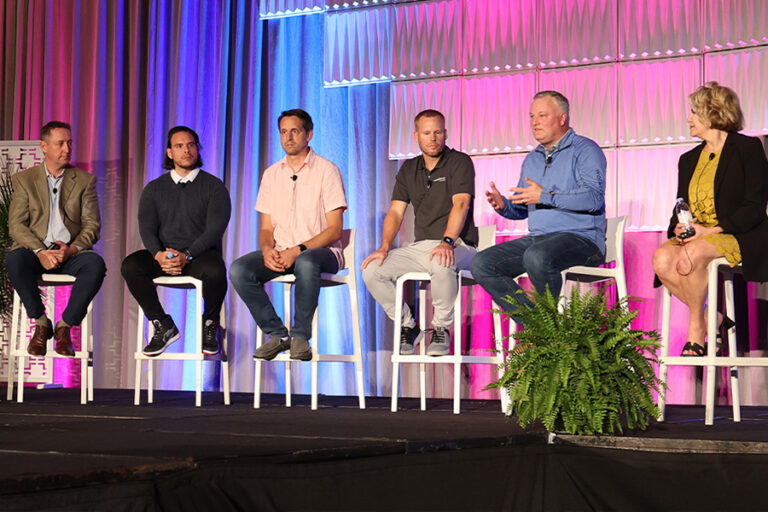 Six people are seated on high stools on a stage, engaged in a panel discussion. The backdrop is lit with blue, purple, and pink lighting. A green plant is placed in front of the stage.