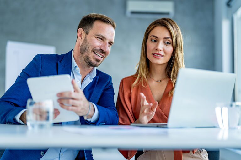 A man in a blue suit holding a tablet and a woman in a brown blouse are sitting at a table, looking at a laptop screen and discussing something.