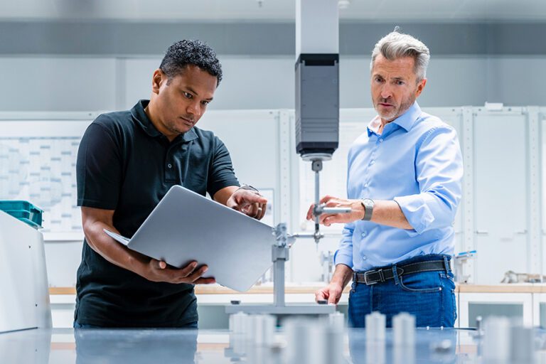 Two men in a laboratory setting, one holding a laptop and pointing, the other observing and adjusting equipment.