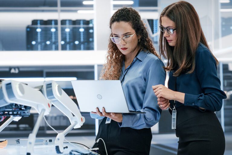 Two women in a modern office setting are examining a laptop screen together. They are wearing formal attire and glasses, and are standing beside tables with various technological equipment.