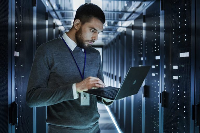 A man holds a laptop while standing in a dimly lit server room. He is wearing a gray sweater, white shirt, and an ID badge.