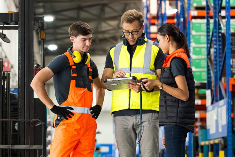 Three warehouse workers review information on a digital tablet in an industrial setting. One is wearing orange overalls, another a high-visibility vest, and the third a dark vest with orange safety trim.